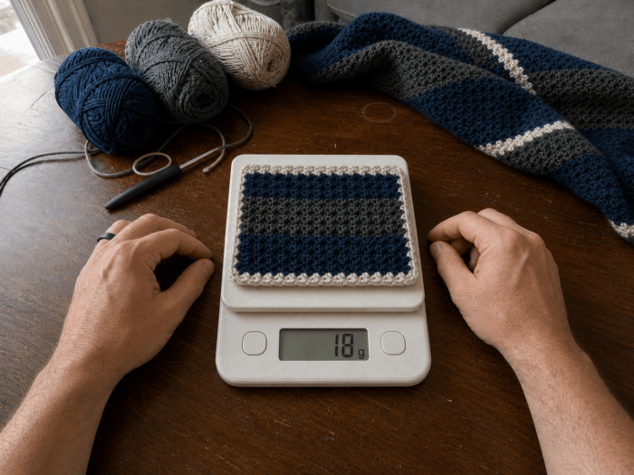 Weighing the matching crochet swatch Estimating yarn needed: A man's bare forearms framing a navy, charcoal, and oatmeal crochet swatch resting on a white digital kitchen scale reading 18 grams, with the matching blanket and yarn nearby.