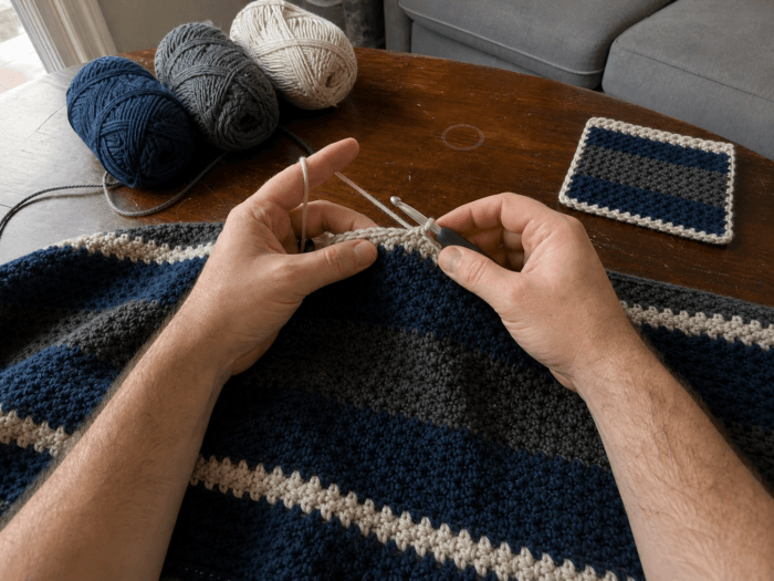 Navy and charcoal crochet blanket project Yarn Yardage Calculator: A man's bare forearms and hands crocheting a navy, charcoal, and oatmeal
throw at a worn walnut table, with matching yarn and a square swatch nearby.